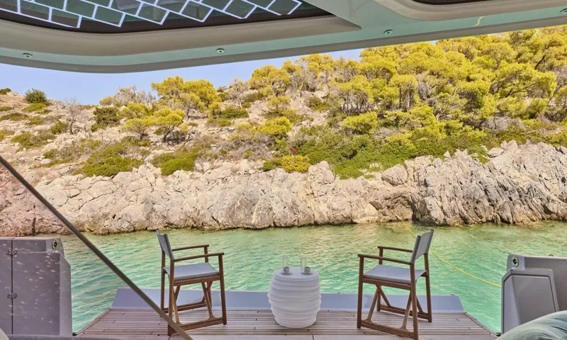 Alina: Two wooden chairs and a small table on a low stern platform facing a rocky green shore