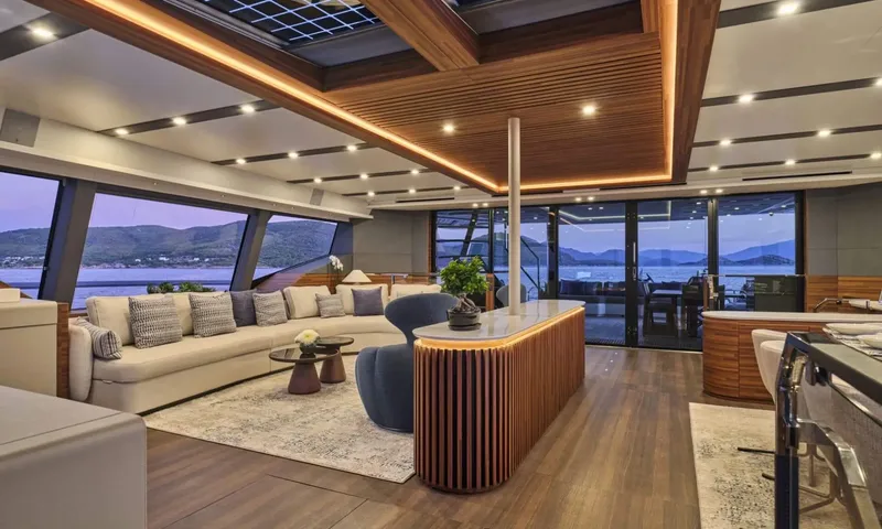 Alina: Open saloon at dusk with cream sofa, wood slatted ceiling, island counter and sea views through large windows