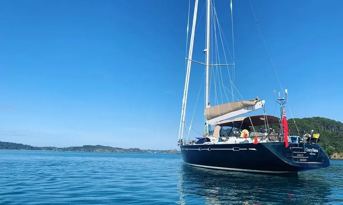 Crazy H: Dark blue hulled sailing yacht at anchor in a calm green bay under clear sky