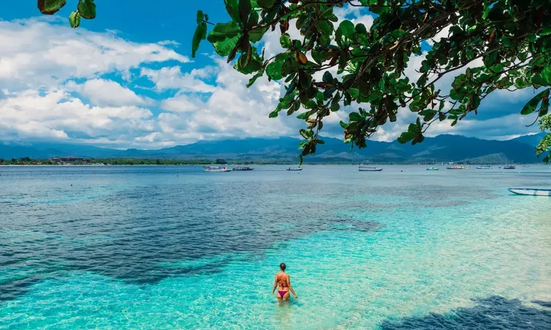 Crazy H: Woman wading in clear turquoise water with moored boats and mountains beyond