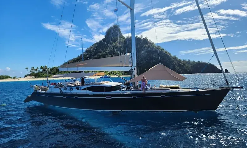 Crazy H: Full side view of dark hulled sailing yacht anchored before a lush island peak
