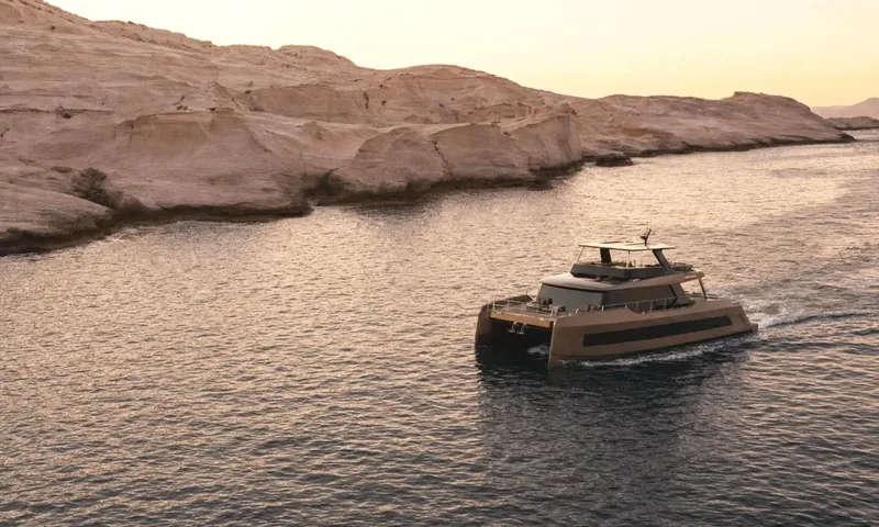 Meltemi: Catamaran cruising past pale rocky cliffs at sunset, seen from above