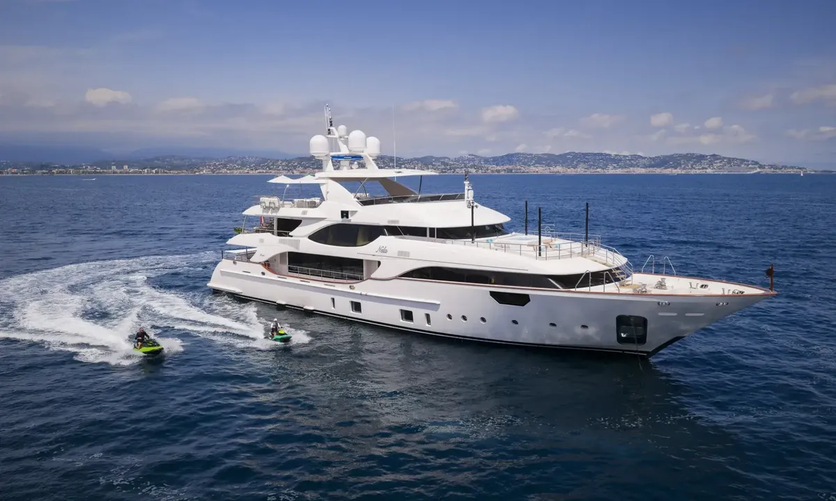 Nela: White yacht cruising on blue sea with two jet skis alongside and a coastal town behind