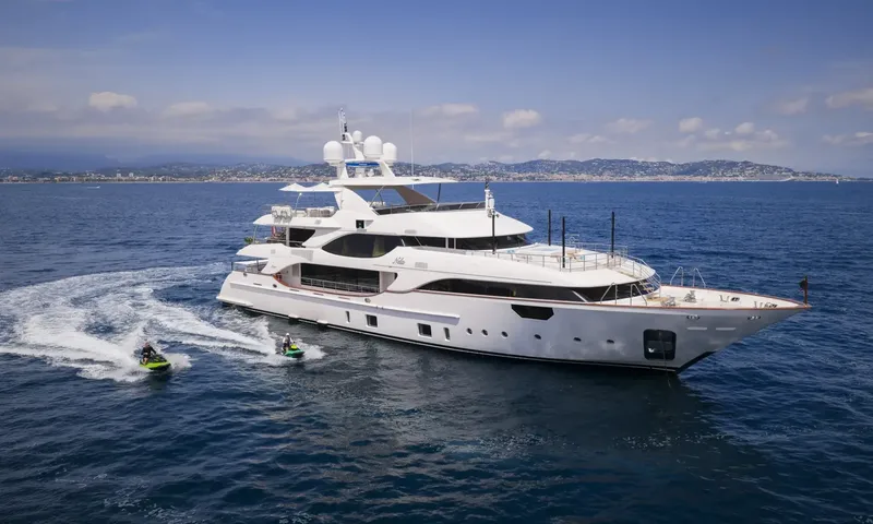 Nela: White yacht cruising on blue sea with two jet skis alongside and a coastal town behind