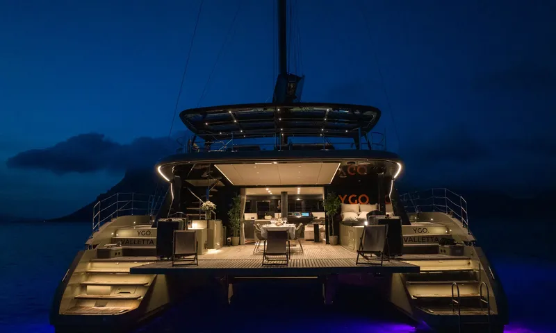YGO: Catamaran stern at night with lit aft deck, dining table and stairs to the water