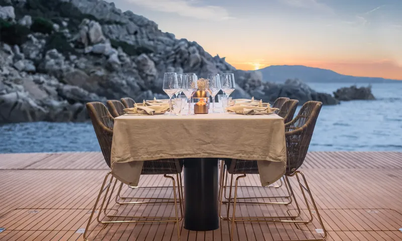 YGO: Table laid for dinner on a teak deck beside rocky coast at sunset