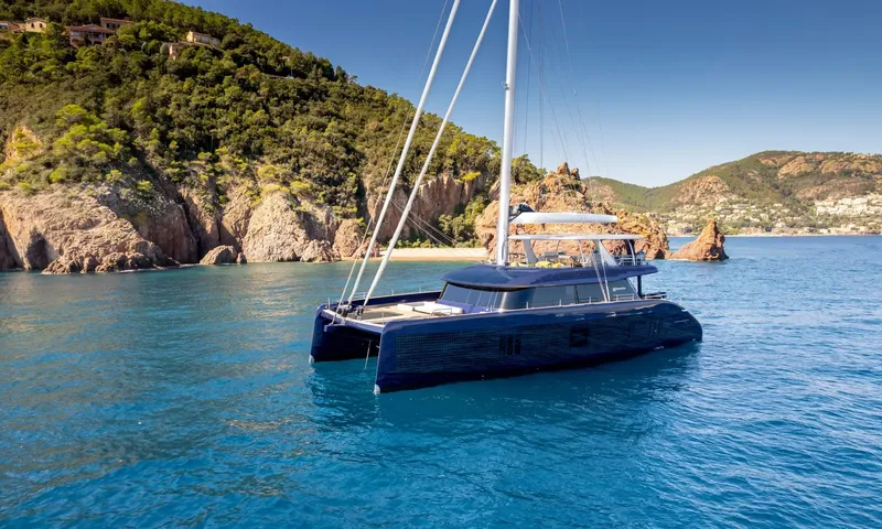 Hu-Lysse: Dark blue sailing catamaran anchored in clear blue water near green hills and rocky shore