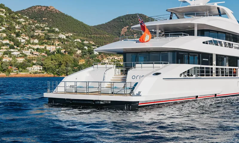 Orion: Stern quarter of a white yacht at anchor with a flag, below a wooded hillside