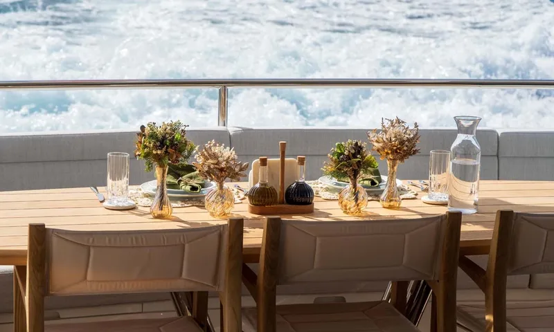 Nigora: Set teak dining table on deck with small vases and churning wake behind